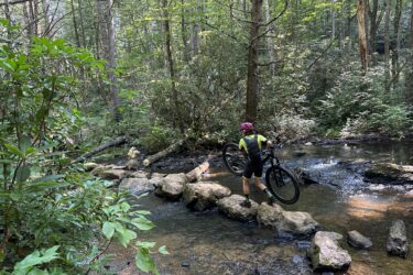 A cyclist navigating a rocky stream in a forested area, carrying a mountain bike over stepping stones while surrounded by lush greenery and sunlight filtering through the trees. Reasonover Creek Trail mountain bike trail.