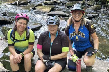 Three women in cycling gear sitting on a rock by a creek, surrounded by greenery. They are wearing helmets and gloves, smiling at the camera. Two women hold water bottles, and they appear to be enjoying a break during a biking outing. Reasonover Creek Trail mountain bike trail.