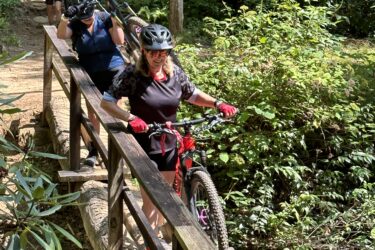 Three cyclists navigate a wooden bridge while carrying their mountain bikes, surrounded by lush greenery and trees on a sunny day. The scene captures the essence of outdoor adventure in a natural setting. Cove Creek Trail / 340 mountain bike trail.