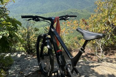 A black mountain bike parked on rocky terrain, overlooking a lush green valley and distant mountains under a clear blue sky. Bright foliage surrounds the scene, adding touches of color to the landscape. Black Mountain mountain bike trail.
