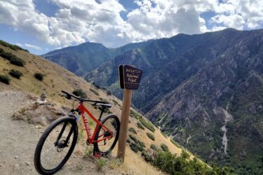 A red mountain bike is leaned against a wooden sign that reads "Wasatch National Forest," set against a backdrop of rolling mountains and partly cloudy skies. The scene captures a trail surrounded by natural beauty, showcasing the outdoors and adventure. Mill Creek Pipeline mountain bike trail.