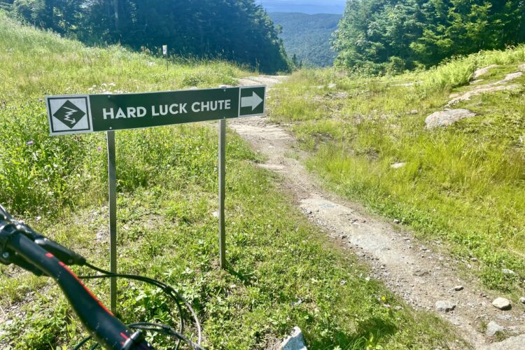 A sign indicating the "Hard Luck Chute" biking trail, positioned next to a dirt path surrounded by greenery and rocky terrain, with mountain views in the background. A bicycle's front fork and handlebars are visible in the foreground.