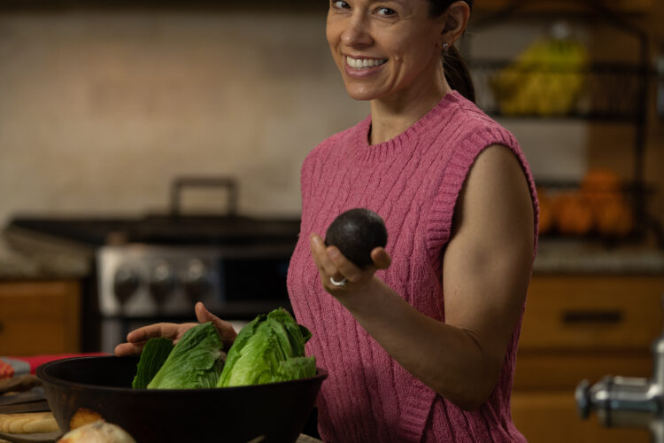 A woman with long dark hair smiles while standing in a kitchen. She is wearing a pink sleeveless sweater and holding an avocado in one hand while an assortment of fresh vegetables, including lettuce and onions, is in a large bowl in front of her. The kitchen features wooden cabinets and a countertop with various other ingredients displayed.