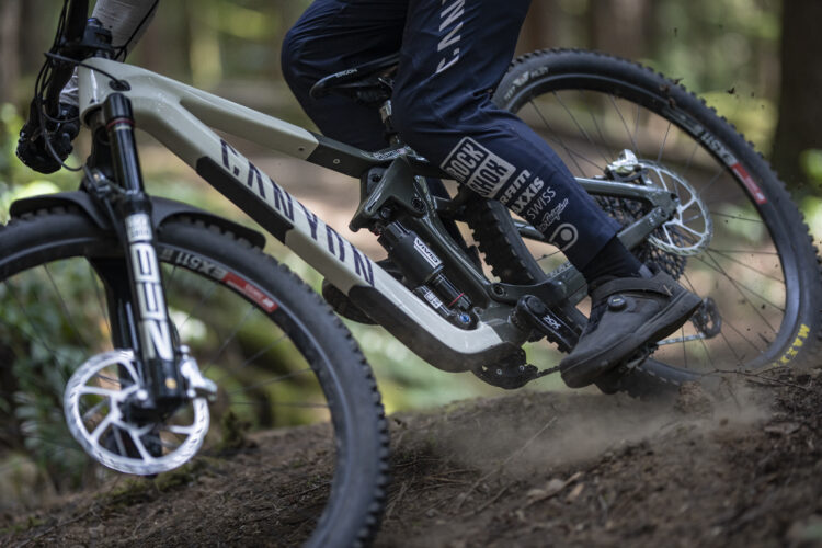 A close-up view of a mountain bike in motion, with a rider's foot positioned on the pedal. The bike has a sleek design and features a suspension system, set against a natural backdrop of trees and dirt. Dust is kicked up from the ground, indicating speed and dynamic movement.