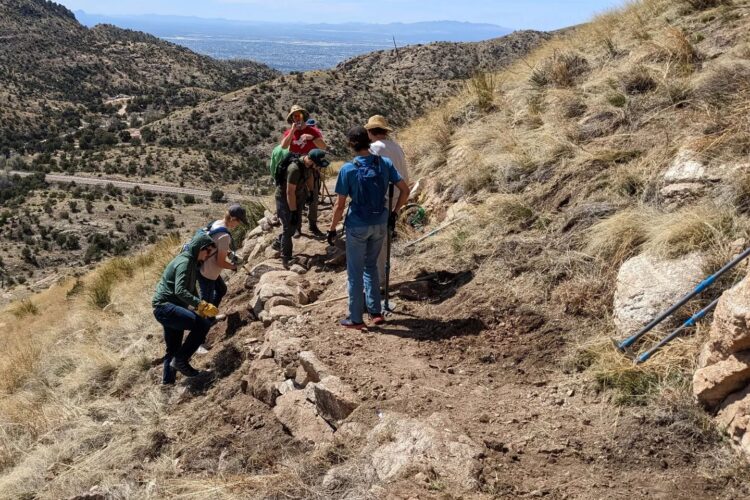 A group of individuals is working on a mountainous trail, actively engaged in trail maintenance. They are scattered along a rocky path surrounded by dry grass and shrubs, with some people using tools to clear debris. The landscape features rolling hills and distant mountains, under a clear blue sky.