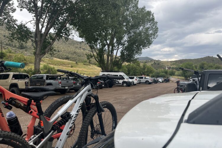 A view of a parking area filled with various vehicles, including trucks and SUVs, surrounded by trees and mountains in the background. Two mountain bikes are prominently displayed in the foreground, resting against a vehicle. The sky is partly cloudy, indicating a potential change in weather.