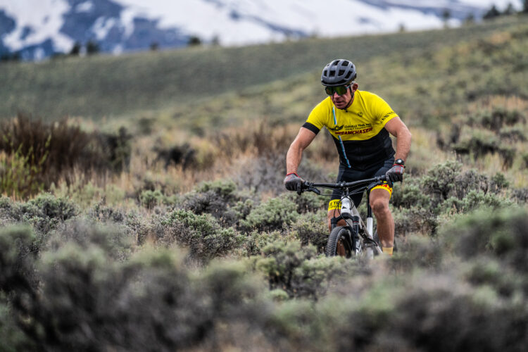 A cyclist dressed in a yellow and black jersey navigates a rocky, shrub-covered trail in a mountainous area, with snow-capped peaks in the background.