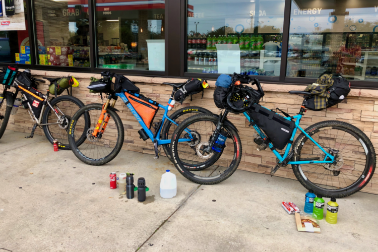 Three bicycles with bikepacking gear parked outside a convenience store, with various water bottles and snacks laid out on the ground. The bicycles are equipped with bags attached to the frames, and a selection of drinks is visible through the store window in the background.