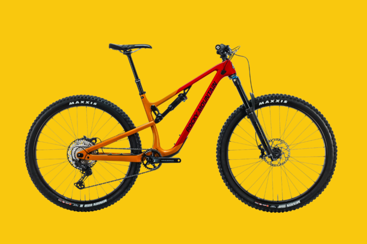 Alt text: A vibrant orange and black Rocky Mountain mountain bike displayed against a bright yellow background. The bike features wide tires with Maxxis branding and a visible drivetrain.