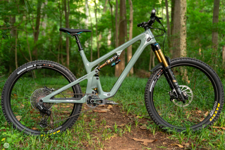 A modern mountain bike with a sleek gray frame, featuring high-performance components and wide tires, is positioned on a grassy patch in a lush, green forest setting. The bike's suspension system is prominently displayed, showcasing its advanced features, while tall trees and greenery create a vibrant natural background.