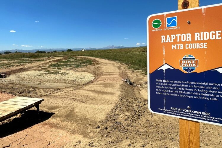 Sign for the Raptor Ridge MTB Course located at a bike park, featuring information on trail difficulty levels (beginner and intermediate). The sign includes safety reminders and describes the skills trails that resemble natural surfaces, emphasizing technical features for mountain biking. The background shows an open field and distant mountains under a clear blue sky.
