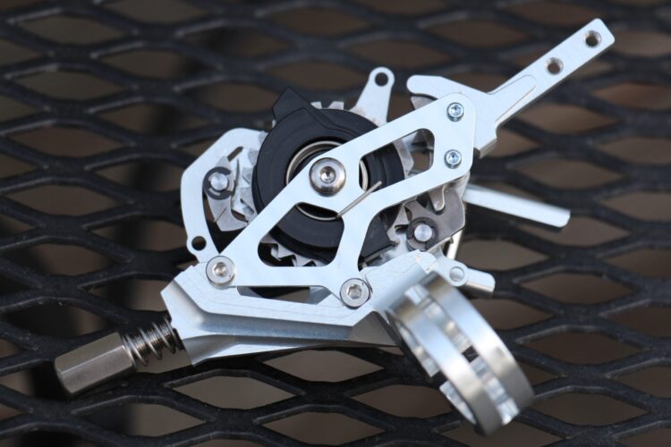A close-up view of a silver mechanical part, possibly a bicycle derailleur or a similar component, set against a black metal grid surface. The part features intricate designs, showcasing gears and levers, with bolts and a sleek metallic finish.