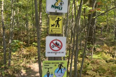 A tall signpost in a forest area, featuring multiple signs. The top sign displays a "WILDCAT" logo, followed by a "THANK YOU!" sign, and a Northern Michigan University logo. Below are several informative signs, including one reminding visitors that dogs must be leashed, a prohibition against motorized vehicles, and a caution about trail conditions for mountain biking. The bottom sign indicates the trail is open for Class 1 e-bikes, with a speed limit of 20 mph and pedal assist only. The surrounding area includes trees and foliage. Noquemanon Trails Network: North Marquette Trails mountain bike trail.