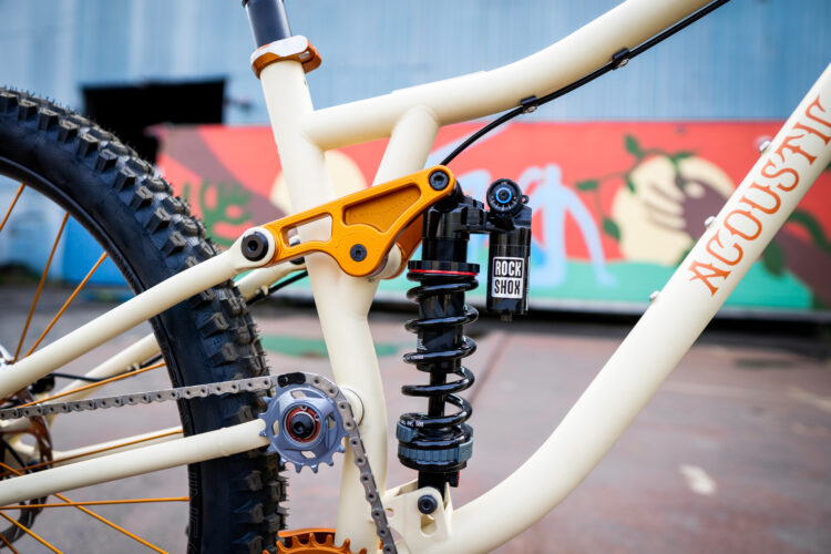 Close-up image of a mountain bike frame featuring a shock absorber and gear system. The frame is primarily cream-colored with an orange accent on the suspension linkage, and a black Rock Shox shock is prominently displayed. The background shows a colorful mural, enhancing the overall dynamic feel of the image.