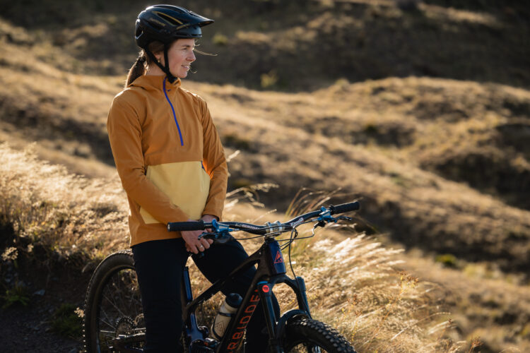 A smiling woman in a brown and yellow cycling jacket and helmet stands next to her mountain bike on a grassy trail. The sun casts a warm glow on the landscape, highlighting the natural surroundings.