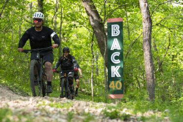 Two mountain bikers riding on a dirt trail surrounded by lush green foliage, with a trail sign reading "BACK 40" prominently displayed nearby. Back 40 mountain bike trail.