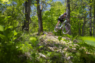 A mountain biker jumps off a trail in a lush, green forest, surrounded by trees and undergrowth. The cyclist is wearing a helmet and riding a mountain bike with vibrant wheels, captured mid-air against a backdrop of clear blue skies and verdant foliage. Hand Cut Hollow mountain bike trail.
