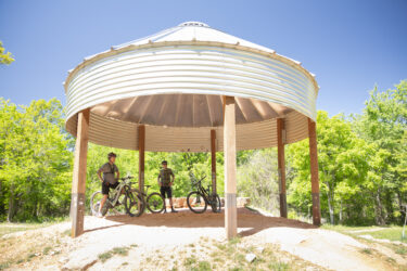 Two mountain bikers stand under a circular metal shelter in a lush green area. They are beside their bikes, enjoying a sunny day. The background features trees and a clear blue sky, creating a vibrant outdoor scene. Hand Cut Hollow mountain bike trail.