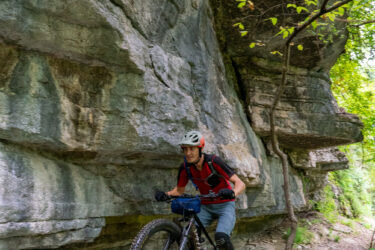 A mountain biker navigates a narrow trail beside a rocky cliff, surrounded by greenery. The cyclist is wearing a helmet and a red shirt, focused on the path ahead. Danger Will Robinson mountain bike trail.