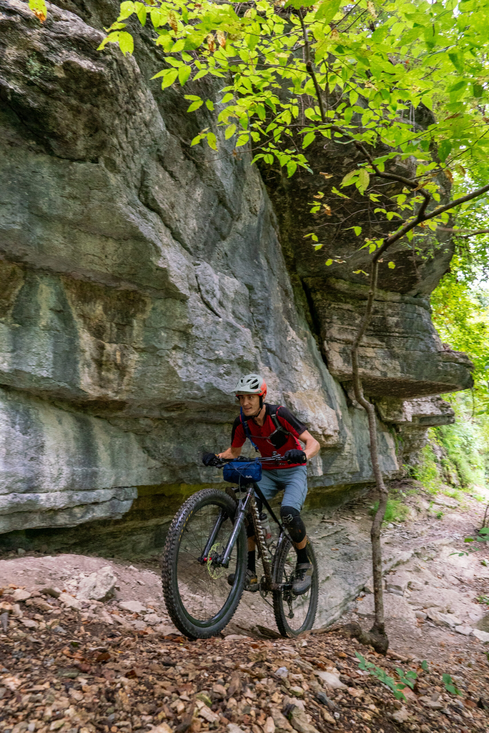 A mountain biker navigates a narrow trail beside a rocky cliff, surrounded by greenery. The cyclist is wearing a helmet and a red shirt, focused on the path ahead. Danger Will Robinson mountain bike trail.