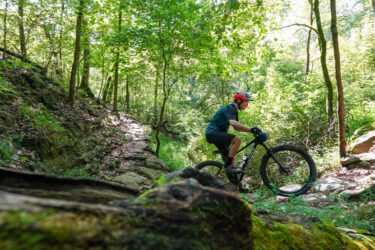 A mountain biker rides along a rocky trail in a lush green forest. Sunlight filters through the trees, illuminating the vibrant foliage and creating a serene outdoor atmosphere. The cyclist wears a helmet and cycling attire, navigating the terrain with focus and skill. Here's Johnny mountain bike trail.