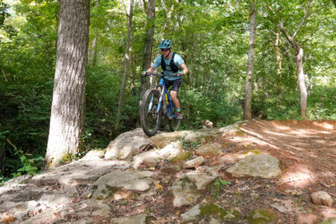 A mountain biker navigating over rocky terrain in a wooded area, with trees and greenery surrounding the trail. The cyclist is wearing a helmet and is focused on the path ahead as they ride their blue mountain bike. Here's Johnny mountain bike trail.
