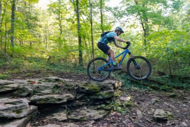 A mountain biker in a blue jersey and helmet leaps off a rocky ledge in a lush green forest. The scene captures the excitement of mountain biking on a natural trail, surrounded by trees and foliage. Oscar's Loop mountain bike trail.