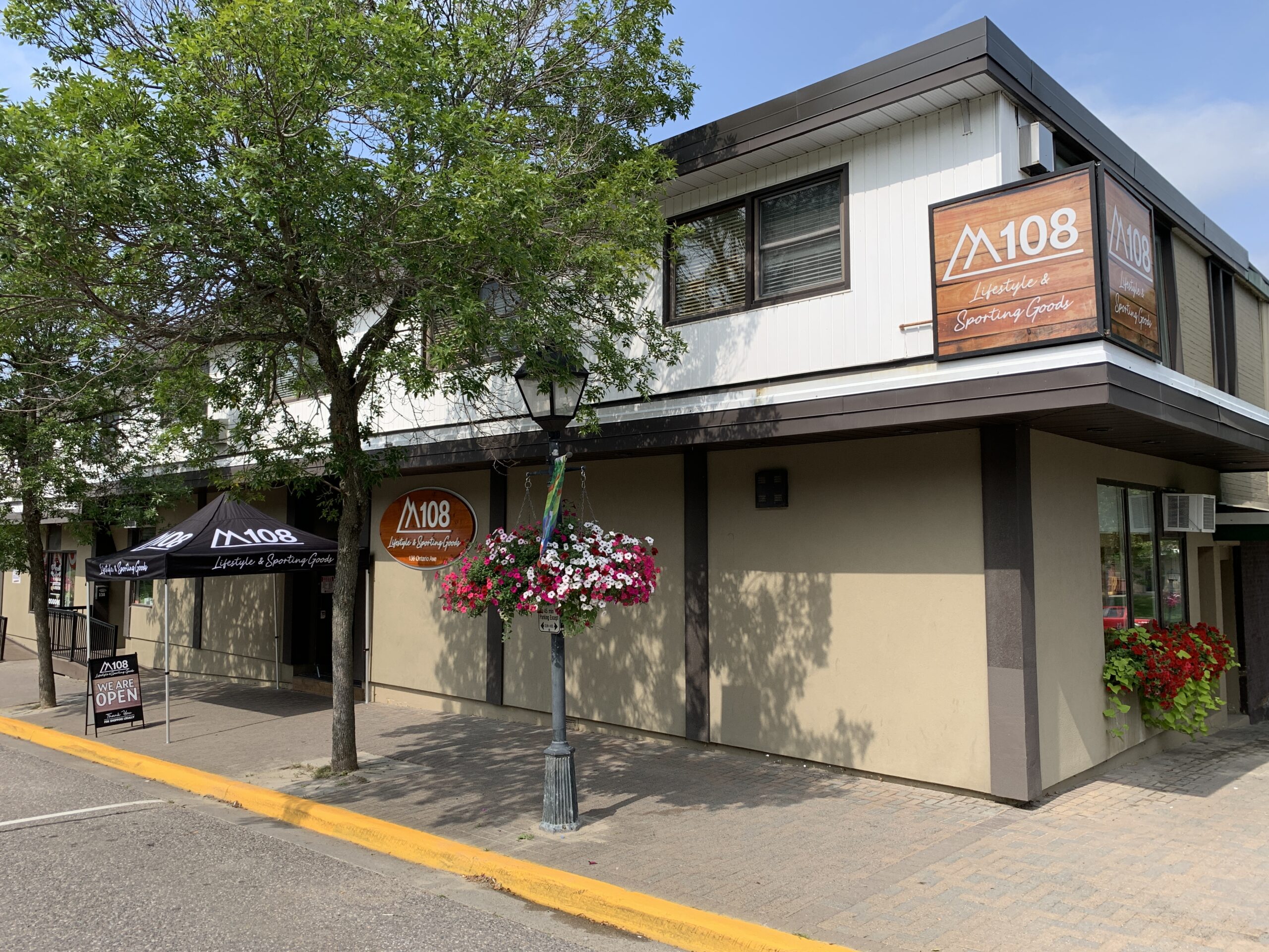 Exterior view of a building featuring the signage "A108 Lifestyle & Sporting Goods." The building has a modern design with large windows and is surrounded by trees and floral decorations. A black tent with the same logo provides coverage at the entrance, and a sign indicates that the store is open. The scene captures a sunny day, enhancing the welcoming atmosphere of the location.