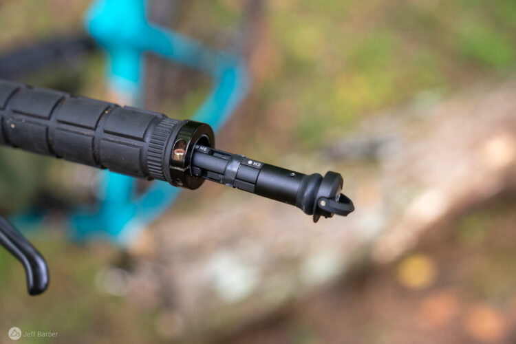 Close-up view of a bicycle handlebar grip featuring a black adjustable lever mechanism. The grip has textured surface for better traction, and the lever is partially extended, showcasing markings for adjustment settings. In the background, blurred outlines of a bicycle frame and natural foliage are visible.