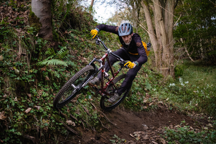 A mountain biker leans into a turn while riding on a wooded trail, surrounded by lush greenery and fallen leaves. The cyclist wears a helmet and gloves, showcasing a focused expression as they navigate the uneven terrain on a red mountain bike.