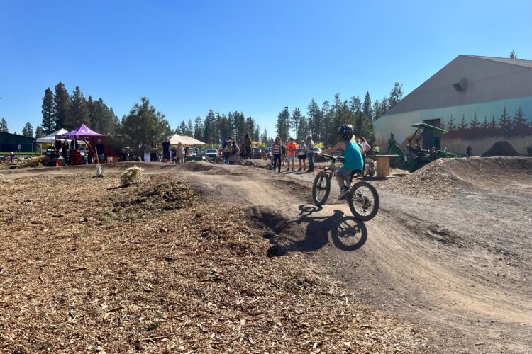 A child wearing a helmet rides a bicycle on a dirt track with jumps, surrounded by spectators and trees against a clear blue sky. In the background, there are tents, vehicles, and a building featuring outdoor murals.