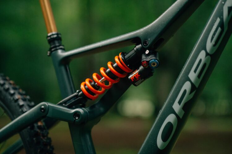 Close-up view of a gray mountain bike frame featuring an orange coil spring suspension and a shock absorber, set against a blurred green forest background.
