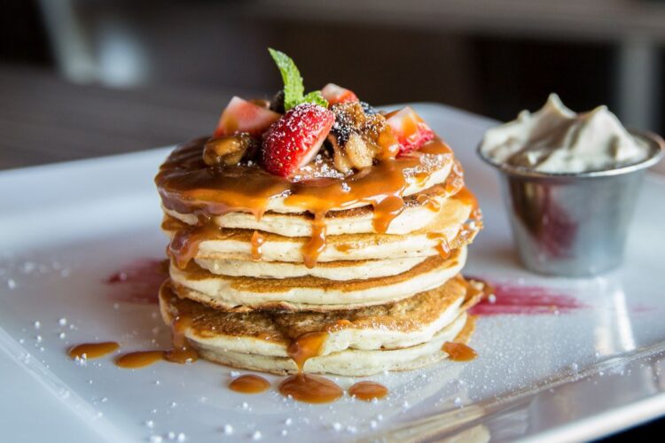 A stack of fluffy pancakes topped with caramel sauce, fresh strawberries, and nuts, garnished with a mint leaf. A small cup of whipped cream is displayed on the side, with a drizzle of sauce on the plate and a dusting of powdered sugar.