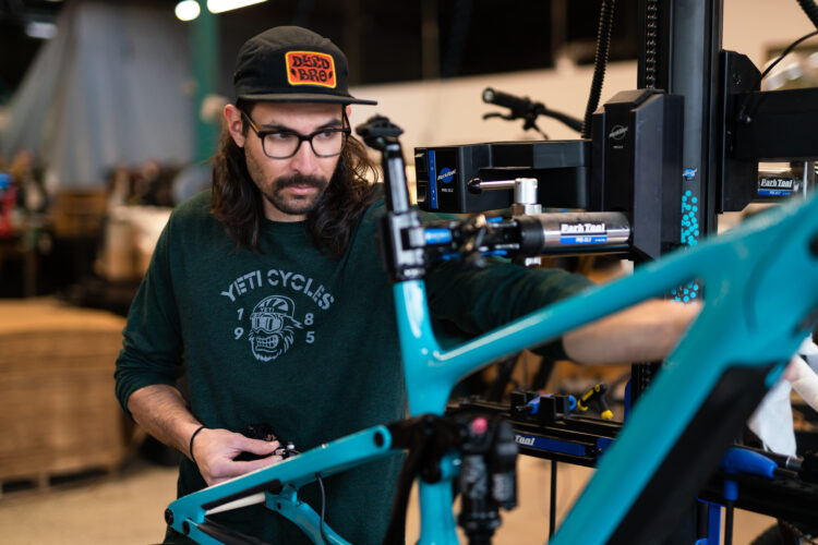 A man with long hair and glasses is working on a bicycle frame in a workshop. He is wearing a green sweatshirt with "YETI CYCLES" printed on it and a black cap. The workspace features various bike repair tools and equipment in the background.