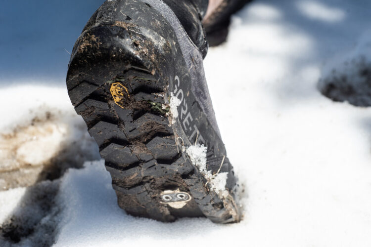 Close-up of a muddy hiking boot with a rugged tread sinking into snow, showcasing dirt, snow, and grass on its surface and highlighting its durable design.