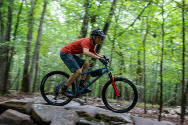A mountain biker in a red shirt and black shorts navigates over rocky terrain in a lush green forest, leaning forward with focused determination, while wearing a helmet and gloves. Southside Park mountain bike trail.