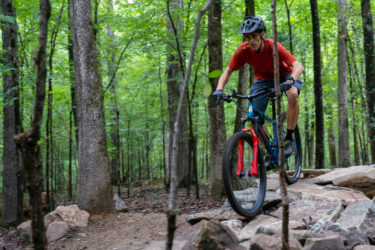 A mountain biker in a red shirt and helmet navigates over rocky terrain on a forest trail, surrounded by lush green trees. Southside Park mountain bike trail.