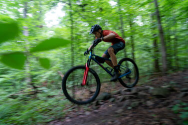 A mountain biker in motion jumps over a rocky section of a forest trail, surrounded by lush green foliage. The image captures a sense of speed and agility as the rider leans forward, wearing a helmet and casual riding gear. Southside Park mountain bike trail.