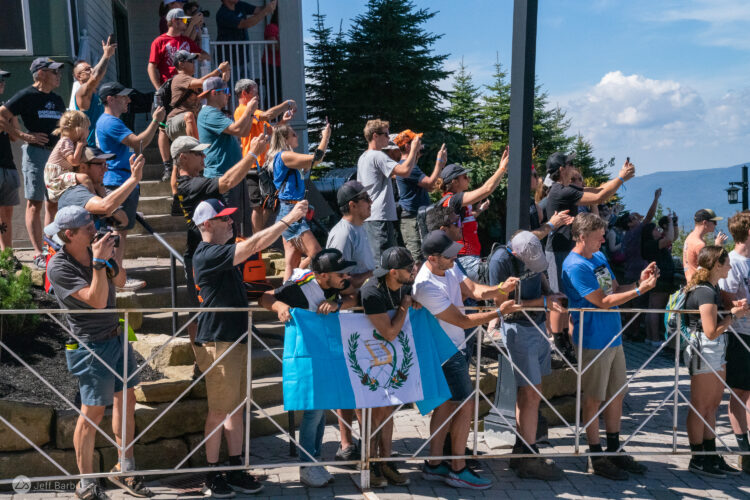 A crowd of excited spectators gathered outdoors, many holding up their phones and cameras to capture the moment. Some individuals are holding a blue and white flag, while others are cheering and enjoying the lively atmosphere. The backdrop features trees and a blue sky, creating a vibrant setting for the event.