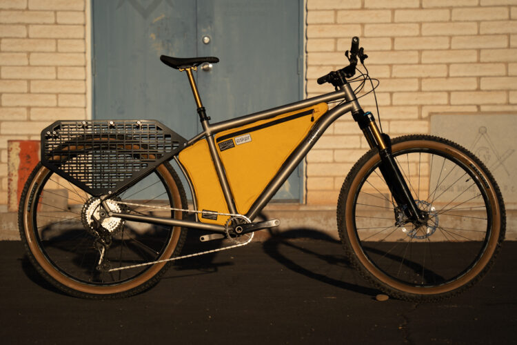 A modern bicycle with a sleek design, featuring a yellow storage bag and a black rear cargo rack. The bike has a gray metallic frame, front suspension, and brown-treaded tires. It is positioned on asphalt in front of a brick wall with a blue door in the background.