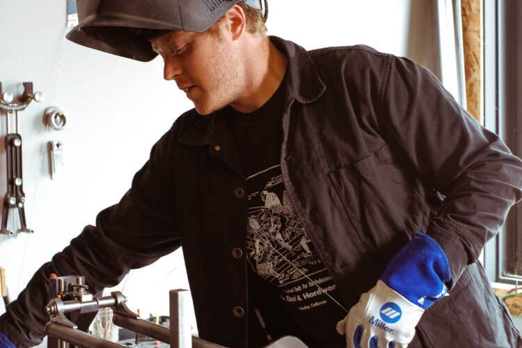 A person wearing a welding helmet and gloves is focused on a workbench, adjusting a metal piece with clamps. The workspace includes various tools visible on the wall in the background, highlighting a hands-on, creative environment.