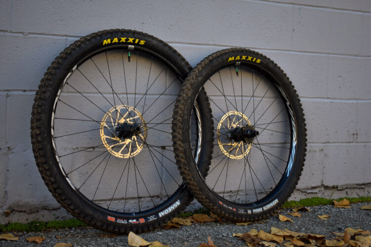 Two mountain bike wheels leaning against a gray wall. Each wheel features knobby Maxxis tires and has a disc brake mounted on the hub. The scene includes fallen leaves on the ground, adding a touch of nature to the urban backdrop.