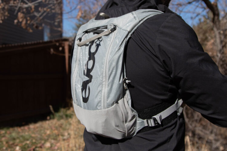 A person wearing a lightweight, gray backpack with a black logo on the back. The setting is outdoors with a blurred background featuring trees and a fence. The individual is dressed in a black jacket, highlighting the backpack's design.