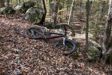 A mountain bike lying on a trail covered with autumn leaves, surrounded by trees and large rocks in a forested area. Voodoo mountain bike trail.