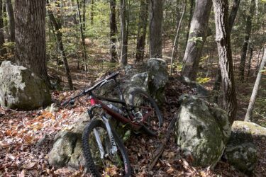 A mountain bike leaning against a large rock, surrounded by autumn leaves and trees in a wooded area. Sunlight filters through the leaves, creating a serene outdoor scene. Voodoo mountain bike trail.