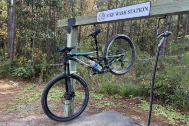 A mountain bike suspended on a bike wash station in a wooded area, with a sign labeled "Bike Wash Station" above it. The bike has colorful accents, and there is a water bottle attached to its frame. Surrounding the station are trees and underbrush, indicating a natural setting. Big Branch Bike Park mountain bike trail.