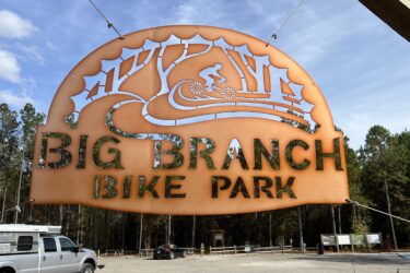 Sign for Big Branch Bike Park, featuring a stylized design of a cyclist and trees, hanging at the park entrance with a natural setting in the background. Big Branch Bike Park mountain bike trail.