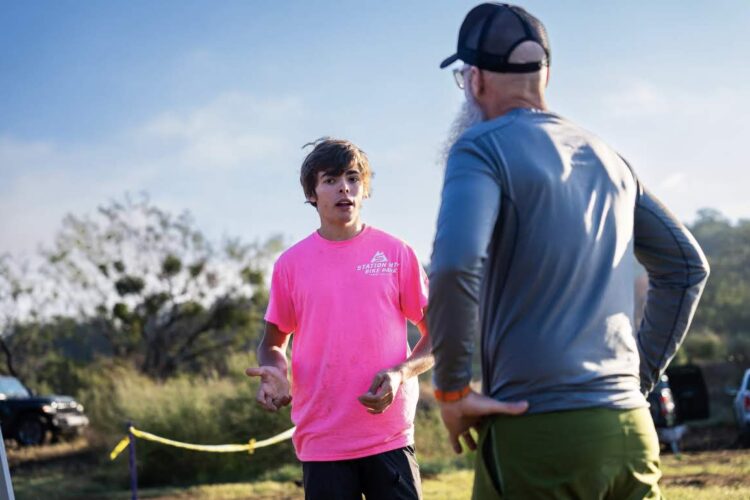 A young man in a bright pink shirt is animatedly speaking to an older man wearing a gray long-sleeve shirt and a black baseball cap. They appear to be in an outdoor setting, with greenery in the background and vehicles visible in the distance. The scene captures a moment of conversation, suggesting a discussion related to an event or activity.