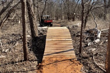 A wooden bridge spans a dirt path in a wooded area, surrounded by bare trees under a clear blue sky. A utility vehicle is partially visible on the left side of the image, indicating the presence of maintenance or construction activity in the natural setting. Wee-chi-ta mountain bike trail.
