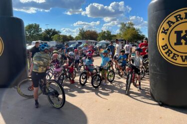 A group of children on bicycles gathers at an outdoor event, excitedly waiting near large inflatable markers. Some children are wearing helmets and colorful clothing, while adults are present in the background. The setting includes blue skies with scattered clouds, trees, and parked vehicles, creating a lively atmosphere for a biking or racing event. Wee-chi-ta mountain bike trail.